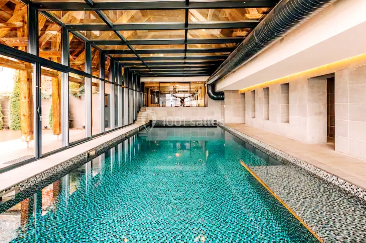 Piscine intérieure avec carrelage mosaïque, entourée de murs en pierre et d'une structure en verre et bois.