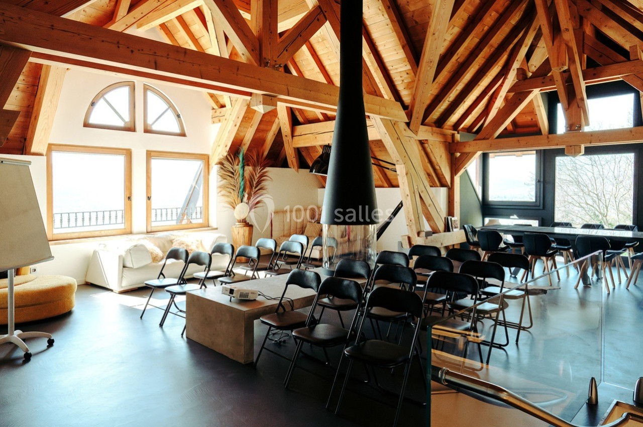 Salle lumineuse avec charpente en bois apparente, aménagée avec des chaises noires disposées face à un tableau blanc.