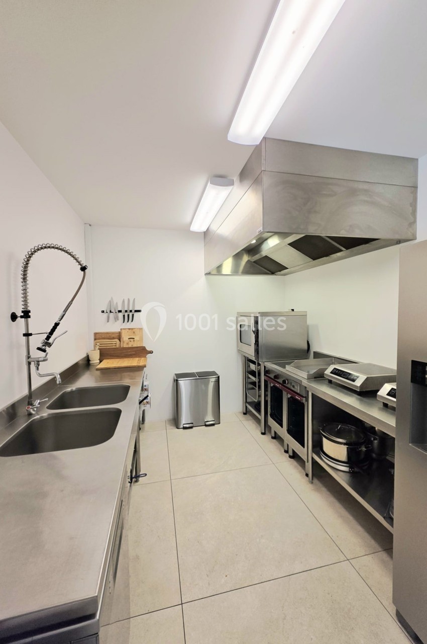 Cuisine professionnelle moderne avec plan de travail en inox, évier, équipements de cuisson et éclairage fluorescent.