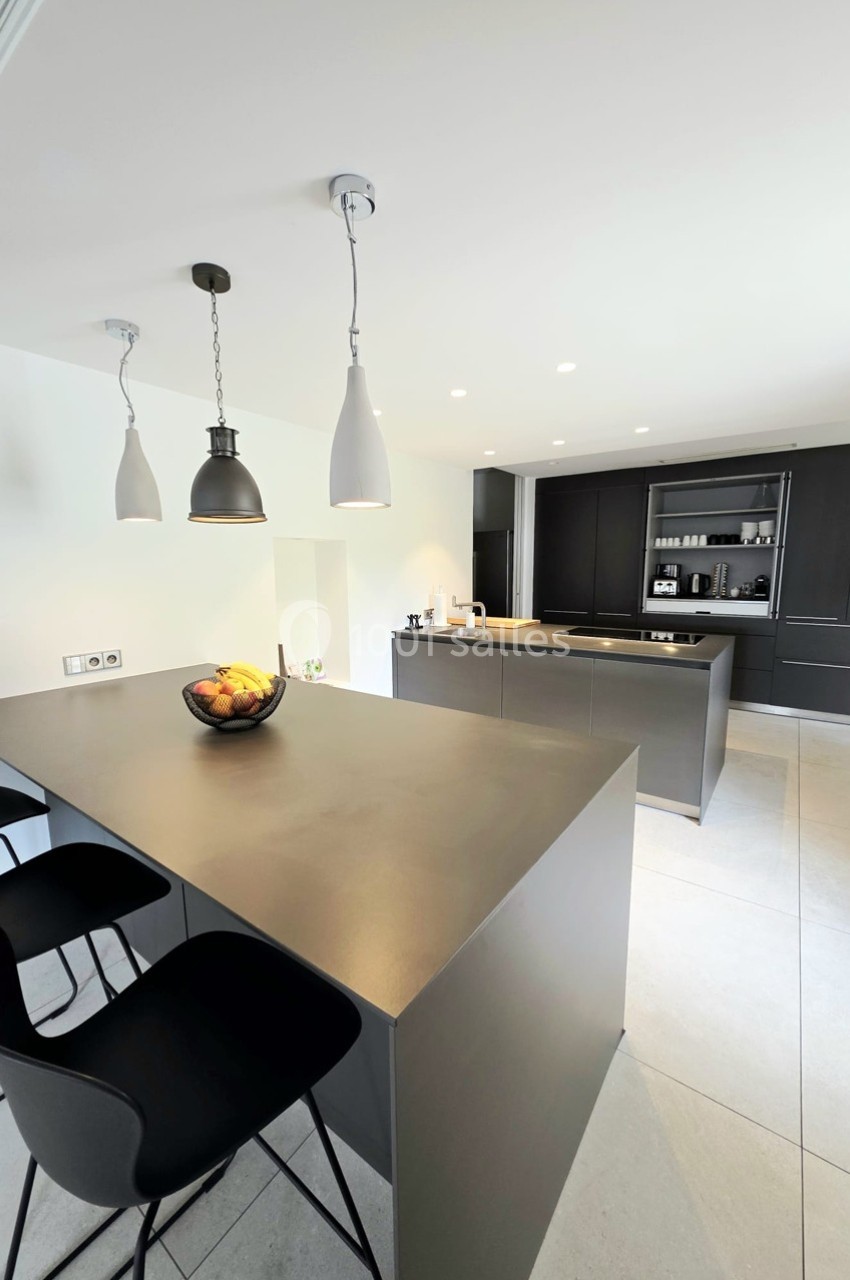 Cuisine moderne avec îlot central noir, plan de travail épuré, chaises hautes et luminaires suspendus.