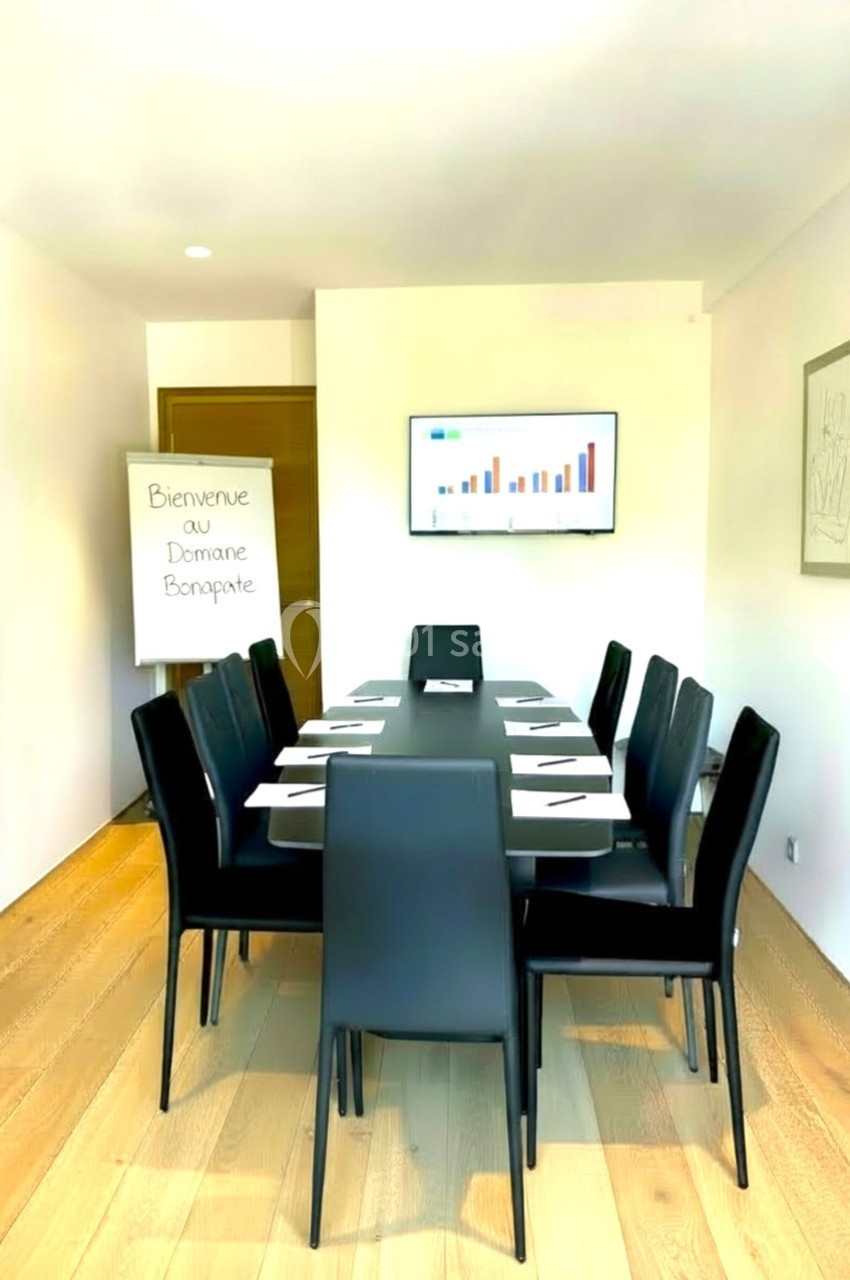 Salle de réunion lumineuse avec table rectangulaire, chaises noires, écran mural et chevalet portant un message de bienvenue.