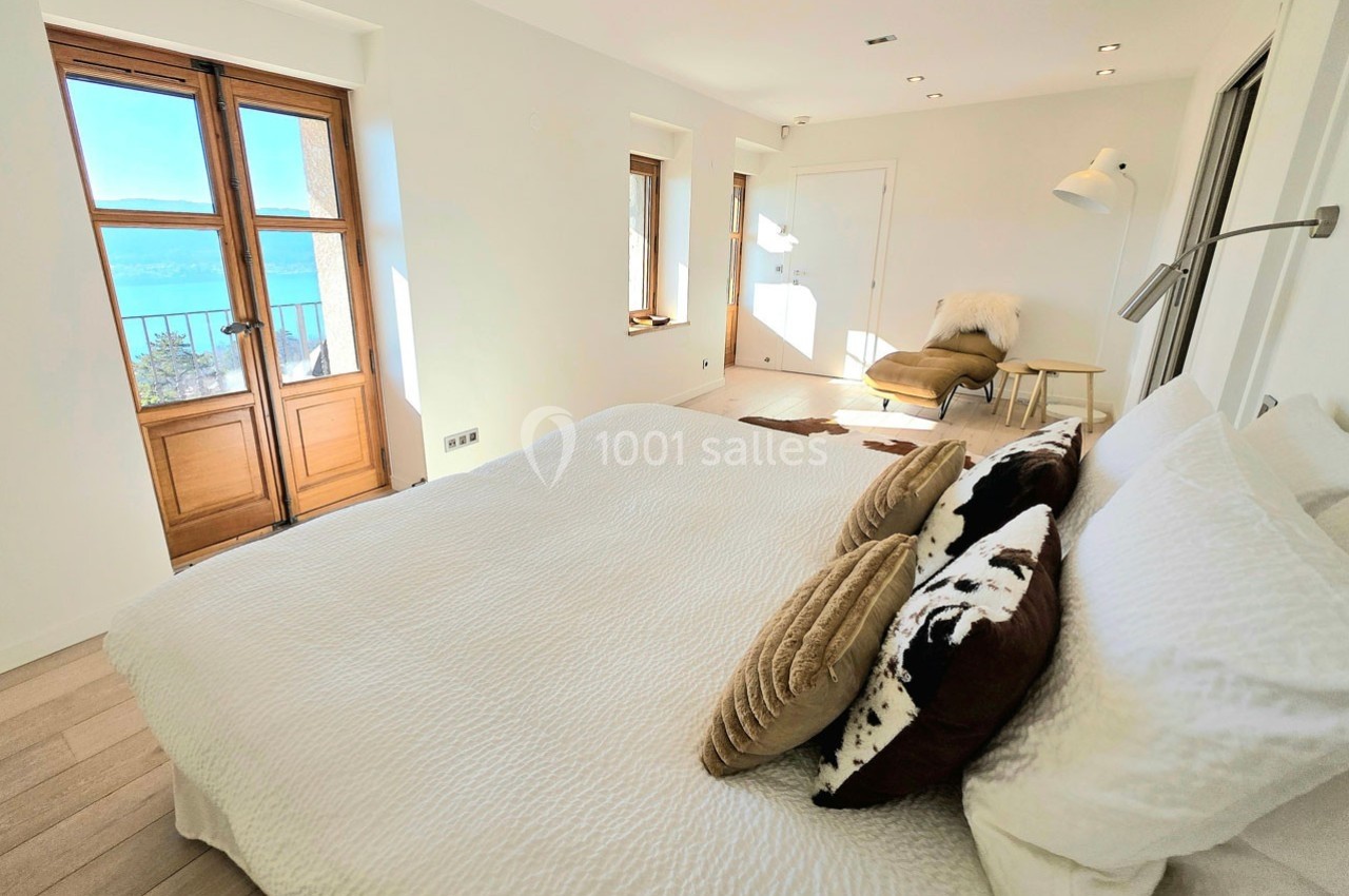 Chambre lumineuse avec lit double, coussins décoratifs, fauteuil et vue sur un balcon donnant sur un paysage ensoleillé.