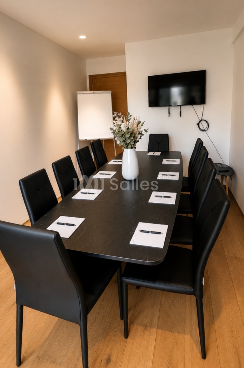 Salle de réunion avec table noire rectangulaire, dix chaises noires, écran mural et projecteur.