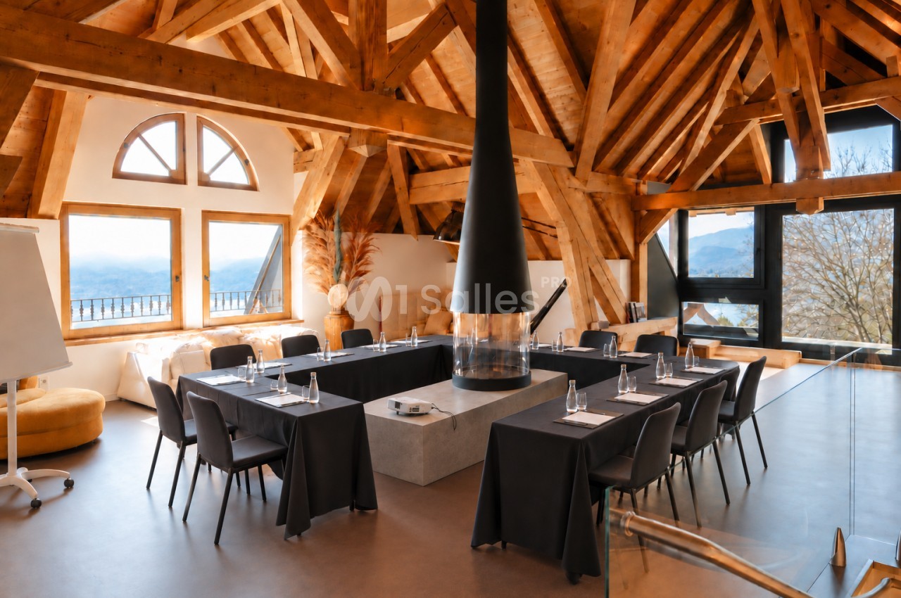 Salle de réunion sous charpente apparente en bois avec tables noires en U, chaises noires et baies vitrées sur montagne.