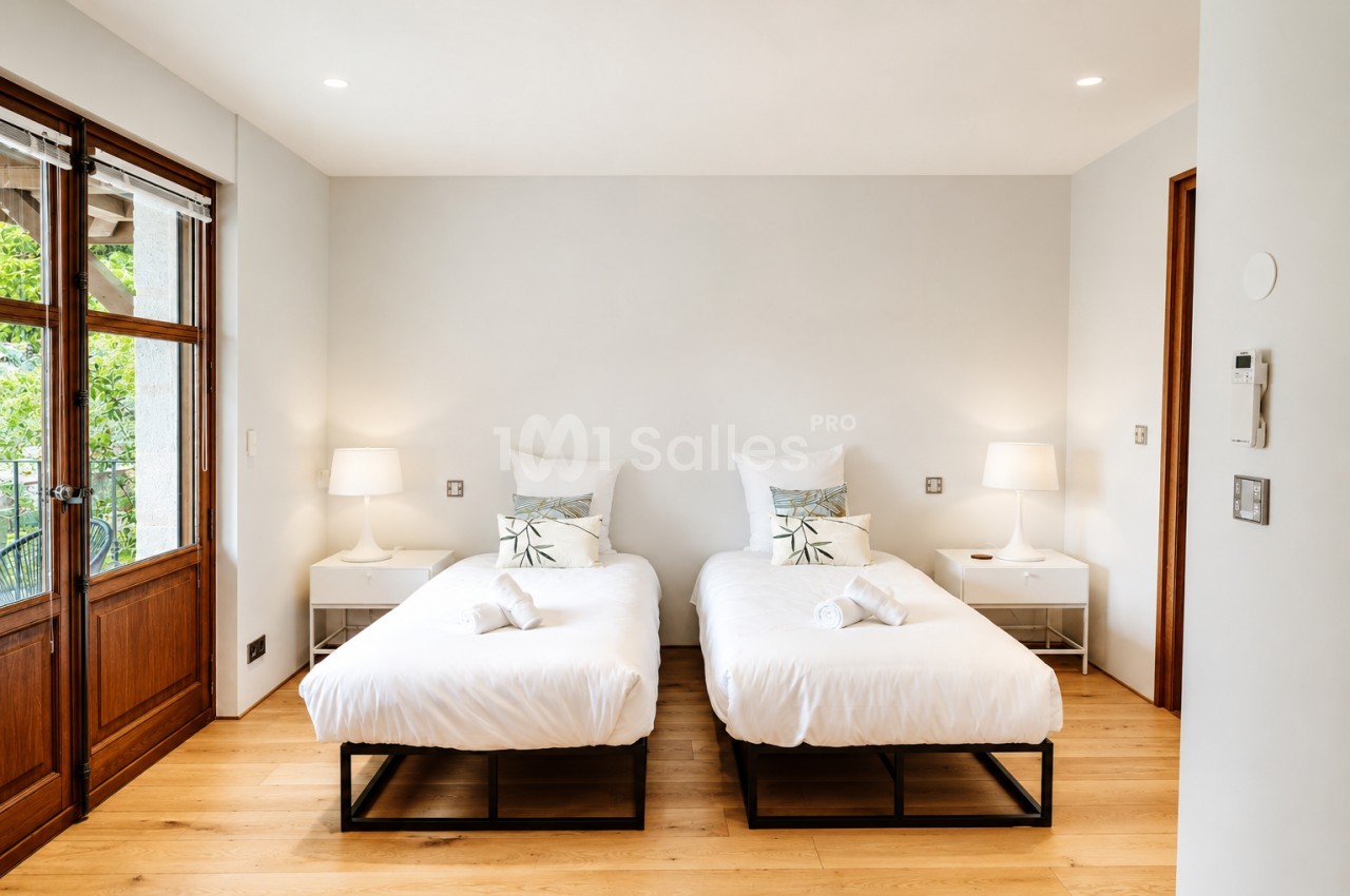 Chambre avec deux lits simples blancs côte à côte, tables de nuit, lampes et portes-fenêtres donnant sur un jardin.