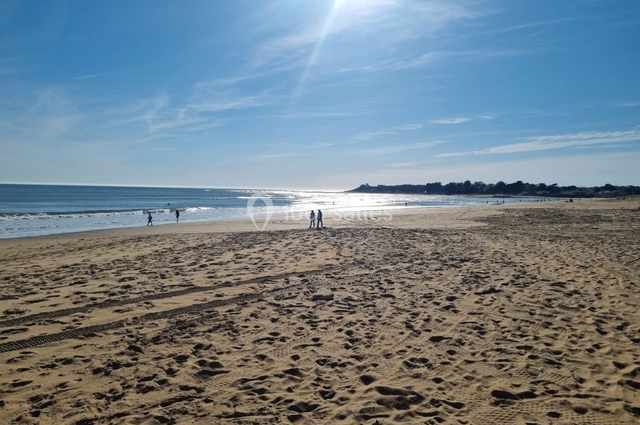 Plage de sable sous un ciel ensoleillé, avec des promeneurs et une vue sur la mer scintillante à l'horizon.