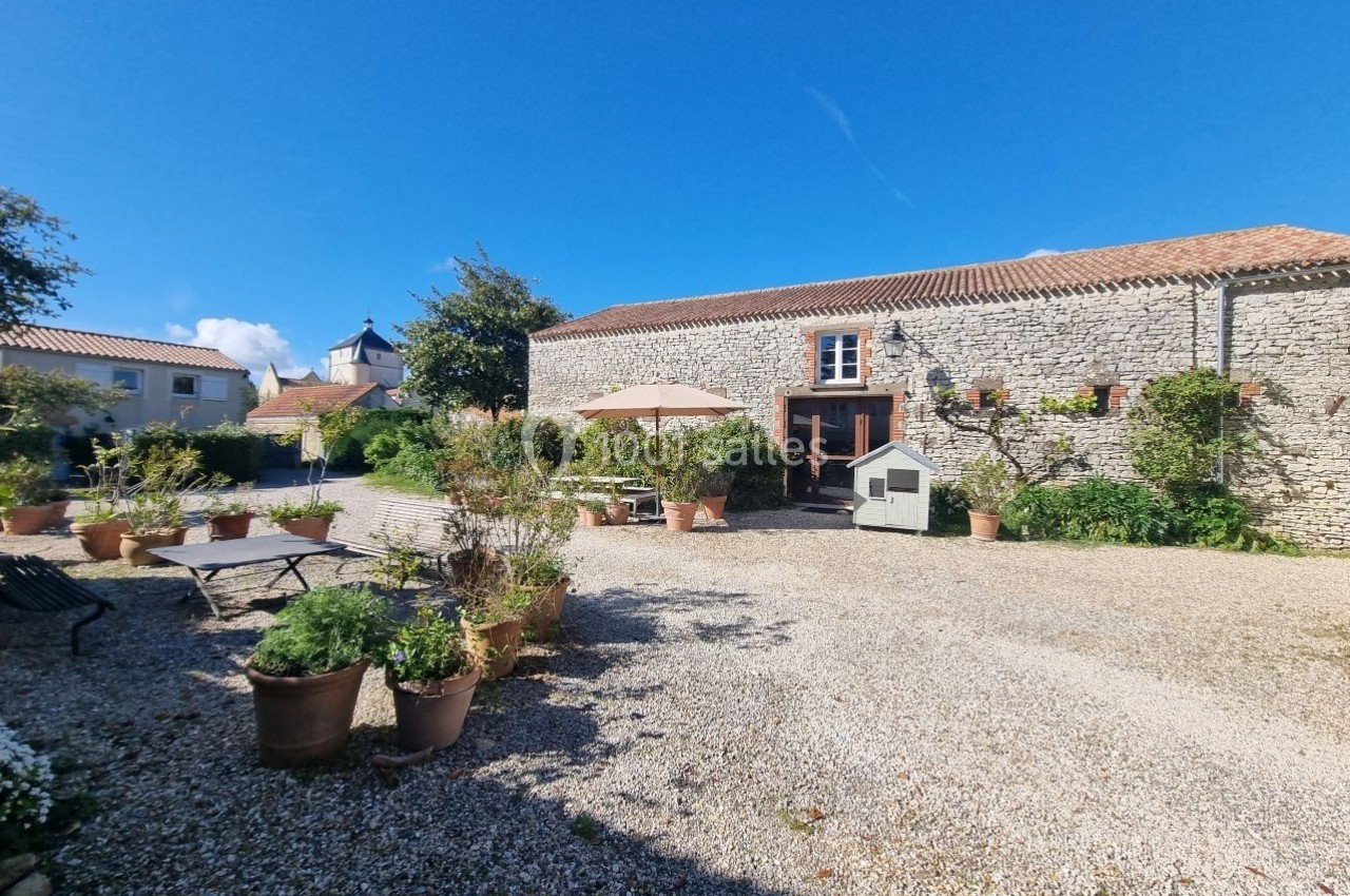 Cour ensoleillée avec graviers, plantes en pots, mobilier de jardin et façade en pierre d'une maison traditionnelle.