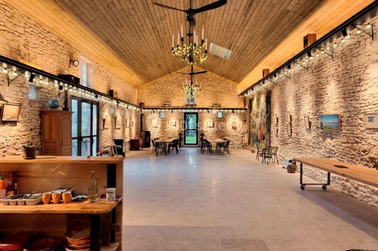 Salle spacieuse avec murs en pierre, plafond en bois, grandes fenêtres et tables disposées pour un événement.