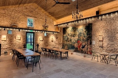 Salle lumineuse avec murs en pierre, grande table en bois, chaises noires et décorations murales, éclairée par des fenêtres.