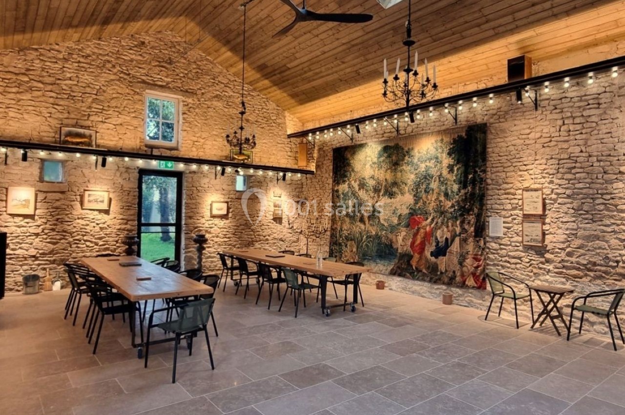 Salle en pierre avec de longues tables en bois, tapisseries murales, lustres et lumière naturelle par des fenêtres.