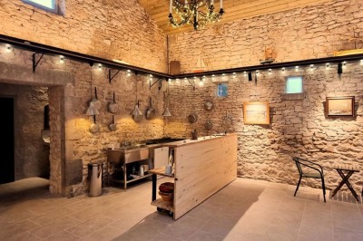 Salle lumineuse avec murs en pierre, grande table en bois, chaises noires et décorations murales, éclairée par des fenêtres.