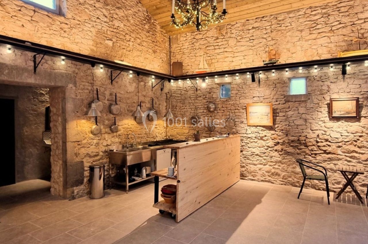 Cuisine rustique avec murs en pierre, éclairage chaleureux, ustensiles accrochés et comptoir en bois clair.