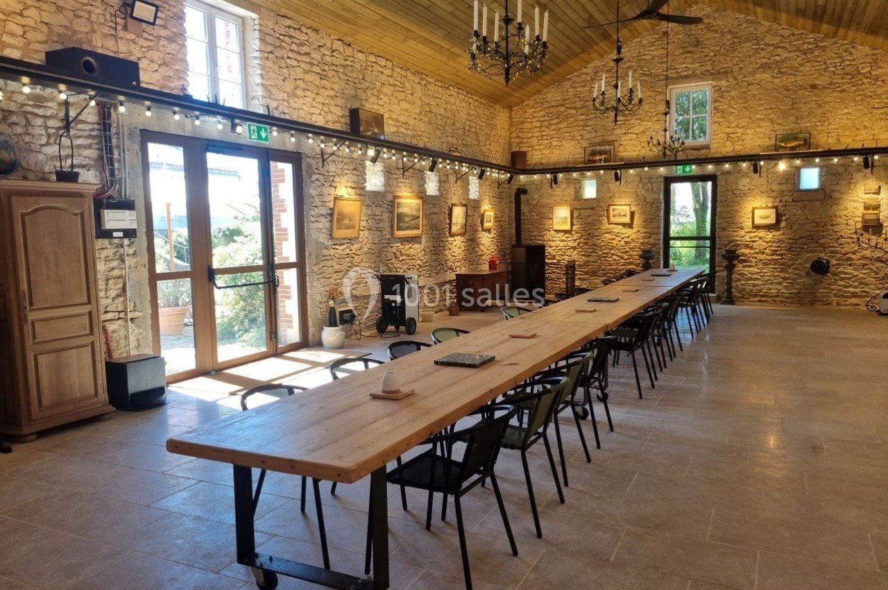 Salle lumineuse avec murs en pierre, grande table en bois, chaises noires et décorations murales, éclairée par des fenêtres.