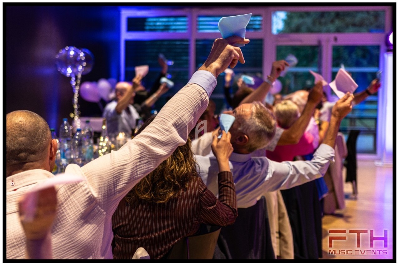 Des convives assis lèvent des pliages en papier coloré dans une salle décorée pour un événement festif.
