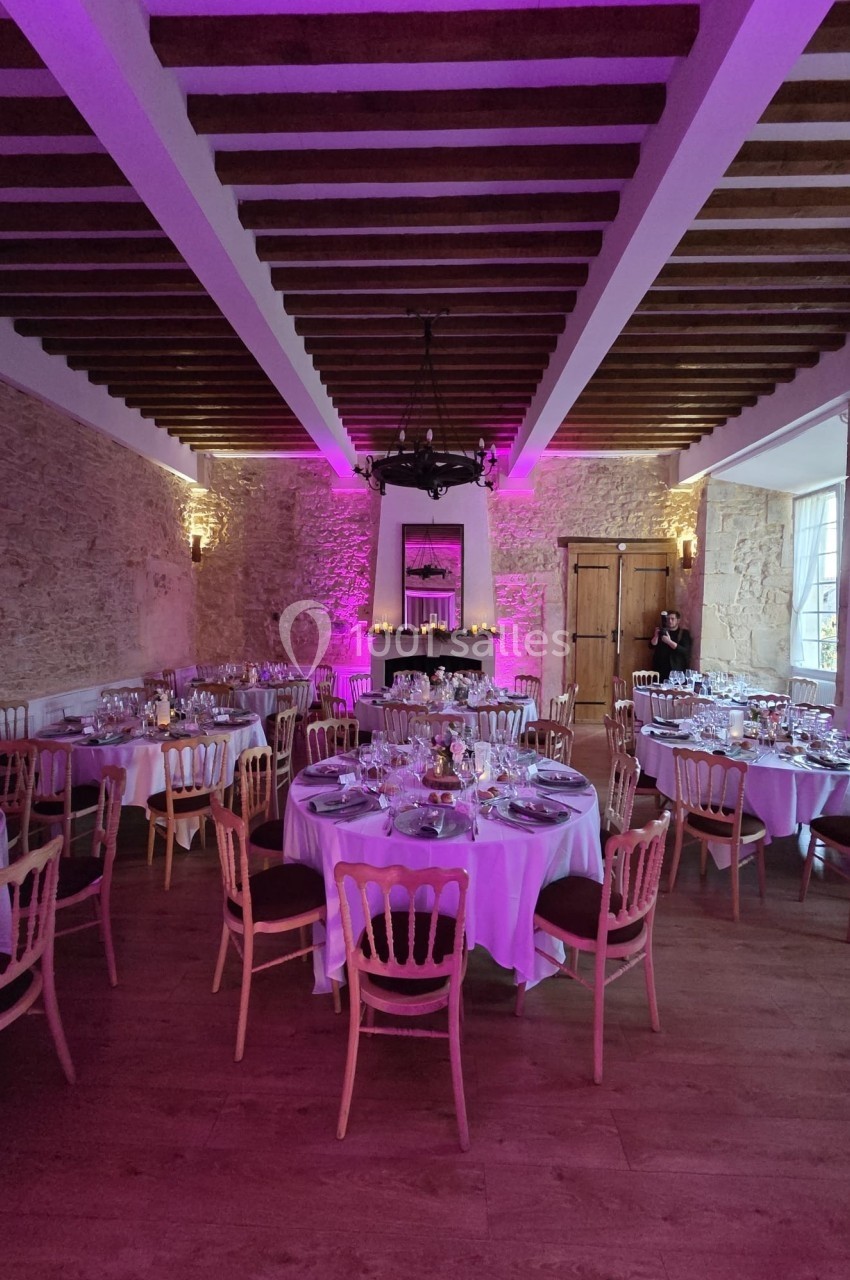 Salle de réception avec tables rondes dressées, éclairage rose et murs en pierre apparente.