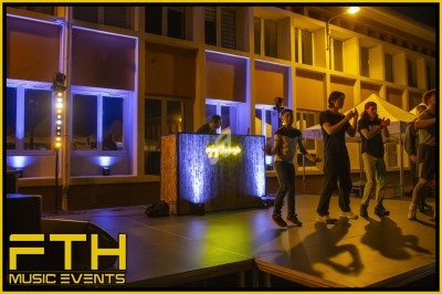 Des personnes sur une scène éclairée applaudissent près d'une table de DJ lumineuse, en extérieur de nuit.
