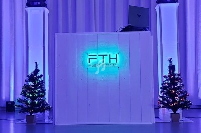 Pupitre de DJ éclairé en bleu avec logo ’FTH Music Events’, entouré de deux sapins décorés et de projecteurs lumineux.