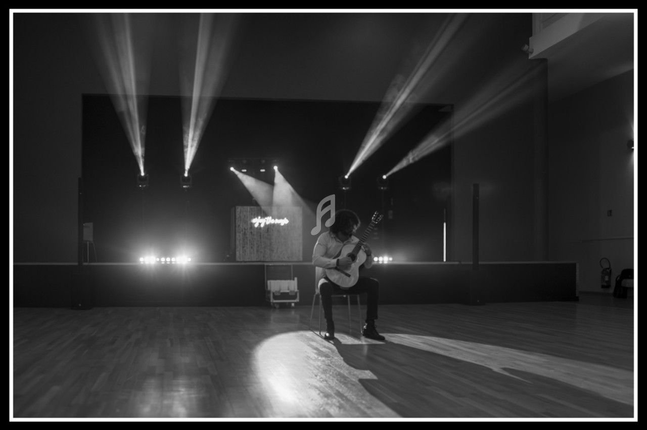 Un musicien joue de la guitare classique sur une scène éclairée par des projecteurs dans une salle sombre.