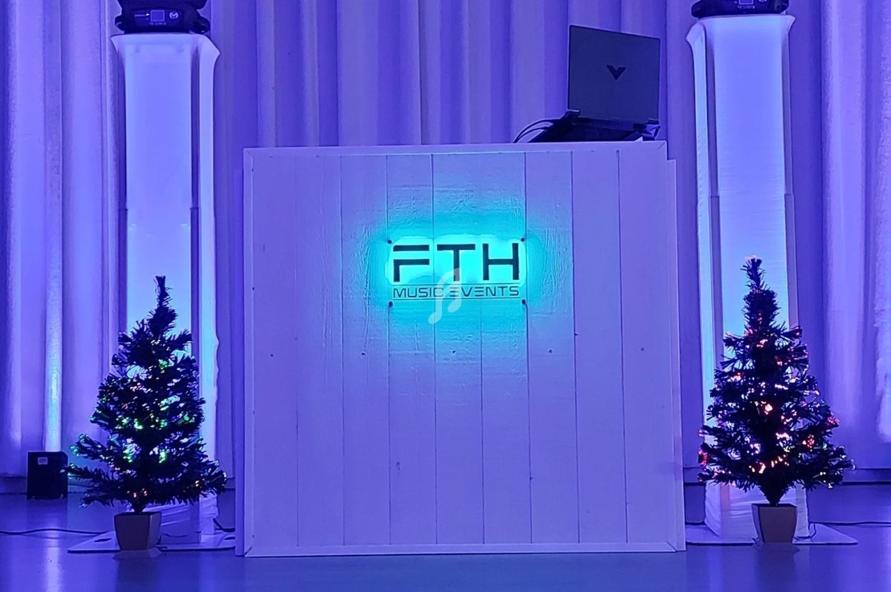 Pupitre de DJ éclairé en bleu avec logo ’FTH Music Events’, entouré de deux sapins décorés et de projecteurs lumineux.