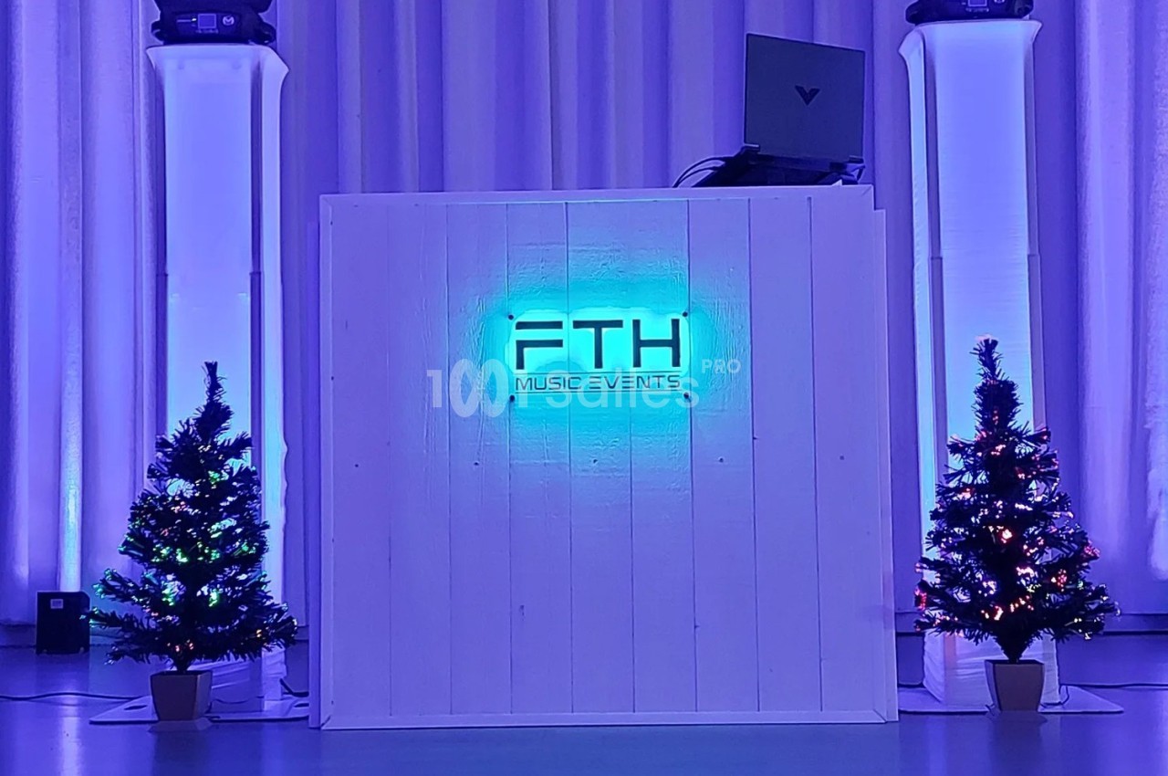 Pupitre de DJ éclairé en bleu avec logo ’FTH Music Events’, entouré de deux sapins décorés et de projecteurs lumineux.