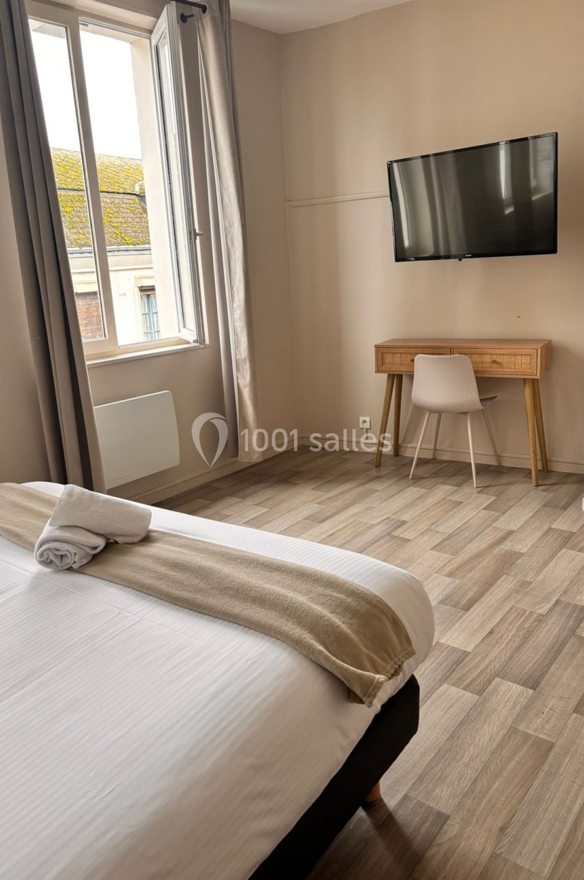 Chambre lumineuse avec lit double, bureau en bois, chaise blanche, télévision murale et fenêtre donnant sur des toits.