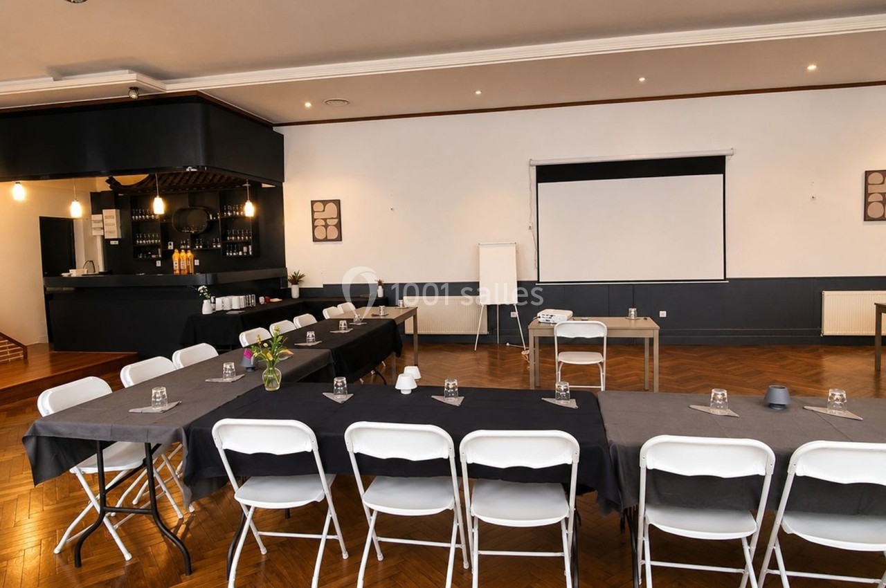 Salle de réunion avec tables disposées en U, chaises blanches, écran de projection et parquet en bois.