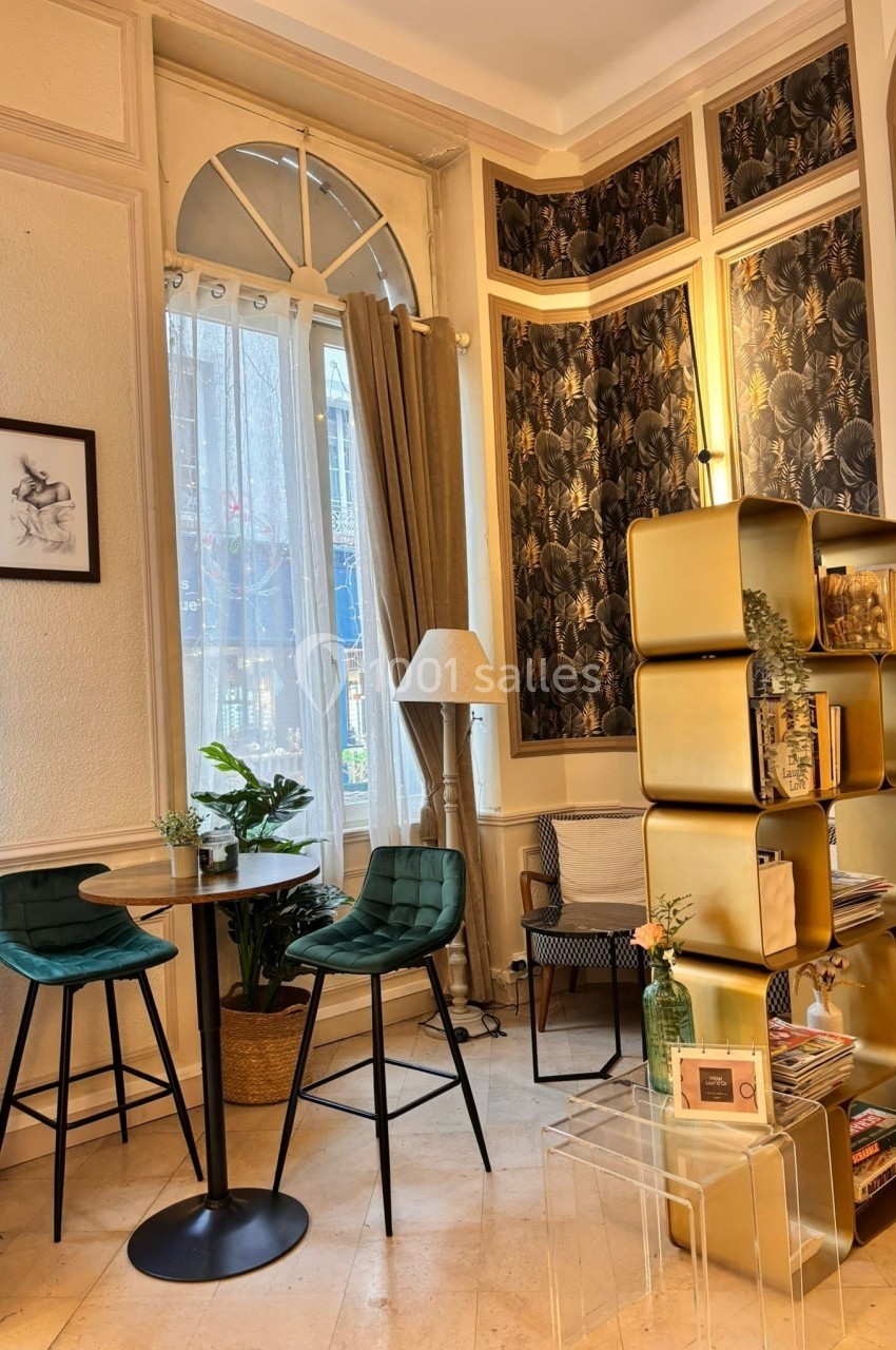 Coin salon lumineux avec table ronde, deux chaises hautes vertes, étagère dorée et plantes près d'une fenêtre.