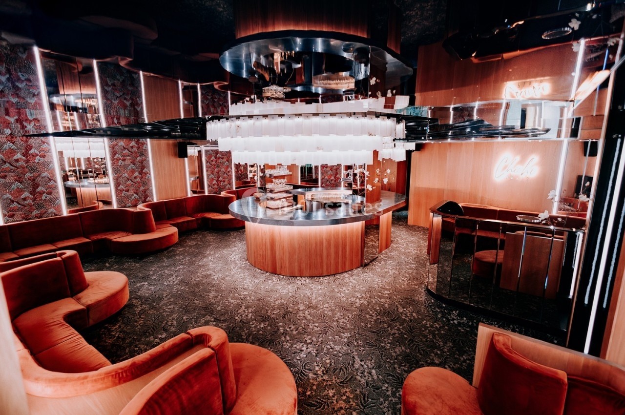 Salon moderne avec banquettes rouges incurvées, bar central circulaire et éclairage suspendu dans une ambiance chaleureuse.