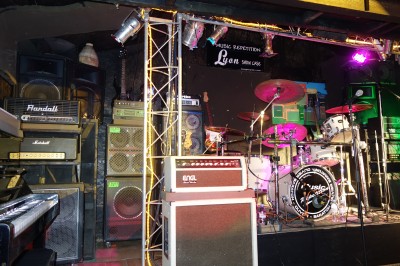 Batterie installée sur une scène éclairée, entourée d'amplificateurs et de structures métalliques.