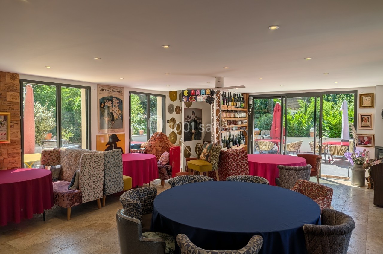 Salle lumineuse avec des tables rondes colorées, des fauteuils variés et de grandes baies vitrées donnant sur un jardin.