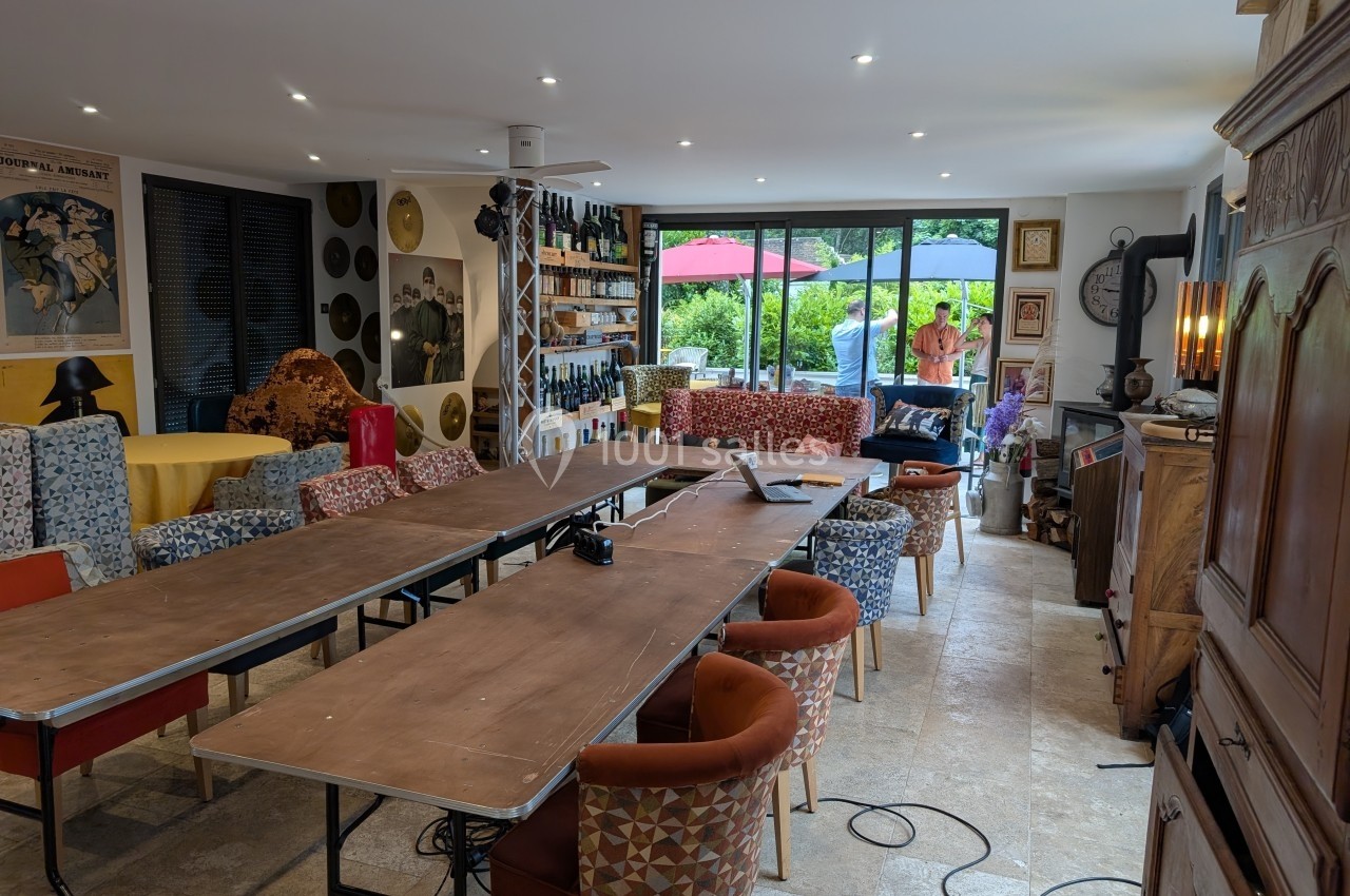 Salle lumineuse avec grande table en bois, chaises colorées, étagères décoratives et vue sur un jardin à travers des baies…
