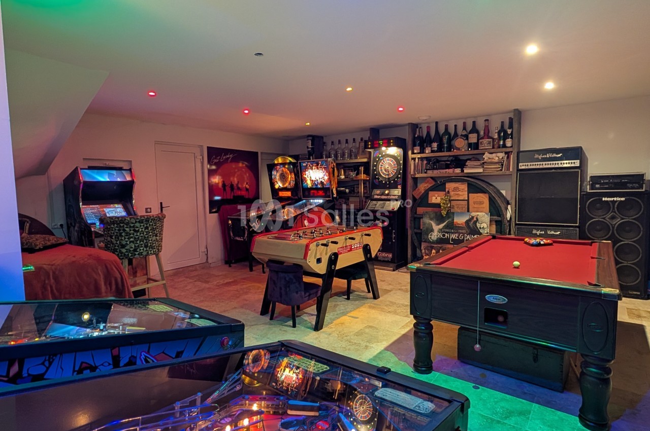 Salle de jeux avec flippers, baby-foot, billard, fléchettes et éclairage coloré dans un espace aménagé.
