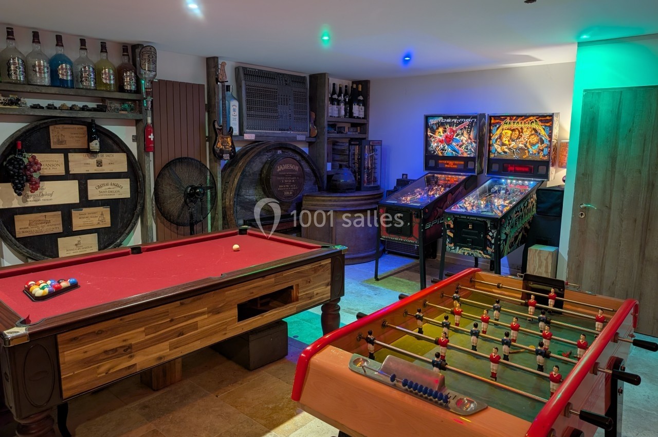 Salle de jeux avec billard, baby-foot, flippers et décorations de bar incluant tonneaux et bouteilles.
