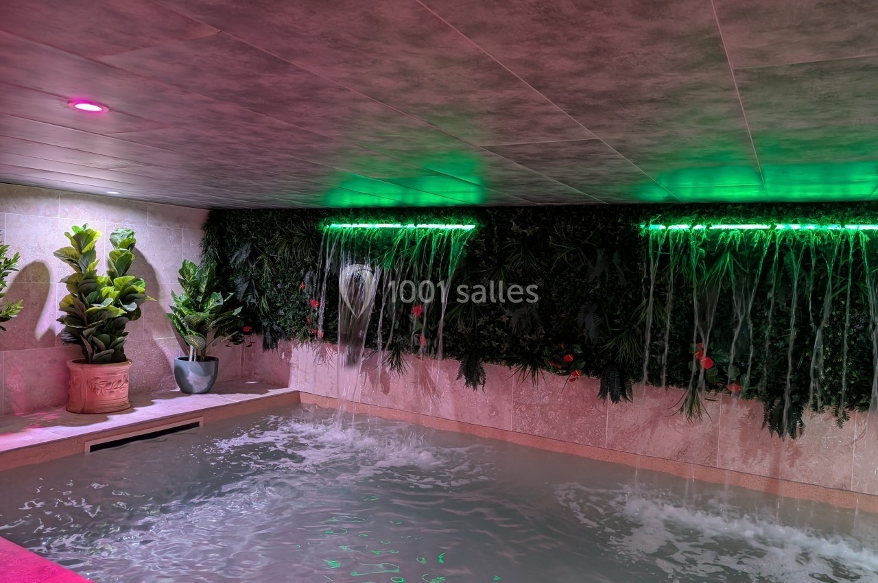 Piscine intérieure avec murs végétalisés, cascades d'eau et éclairage tamisé rose et vert.