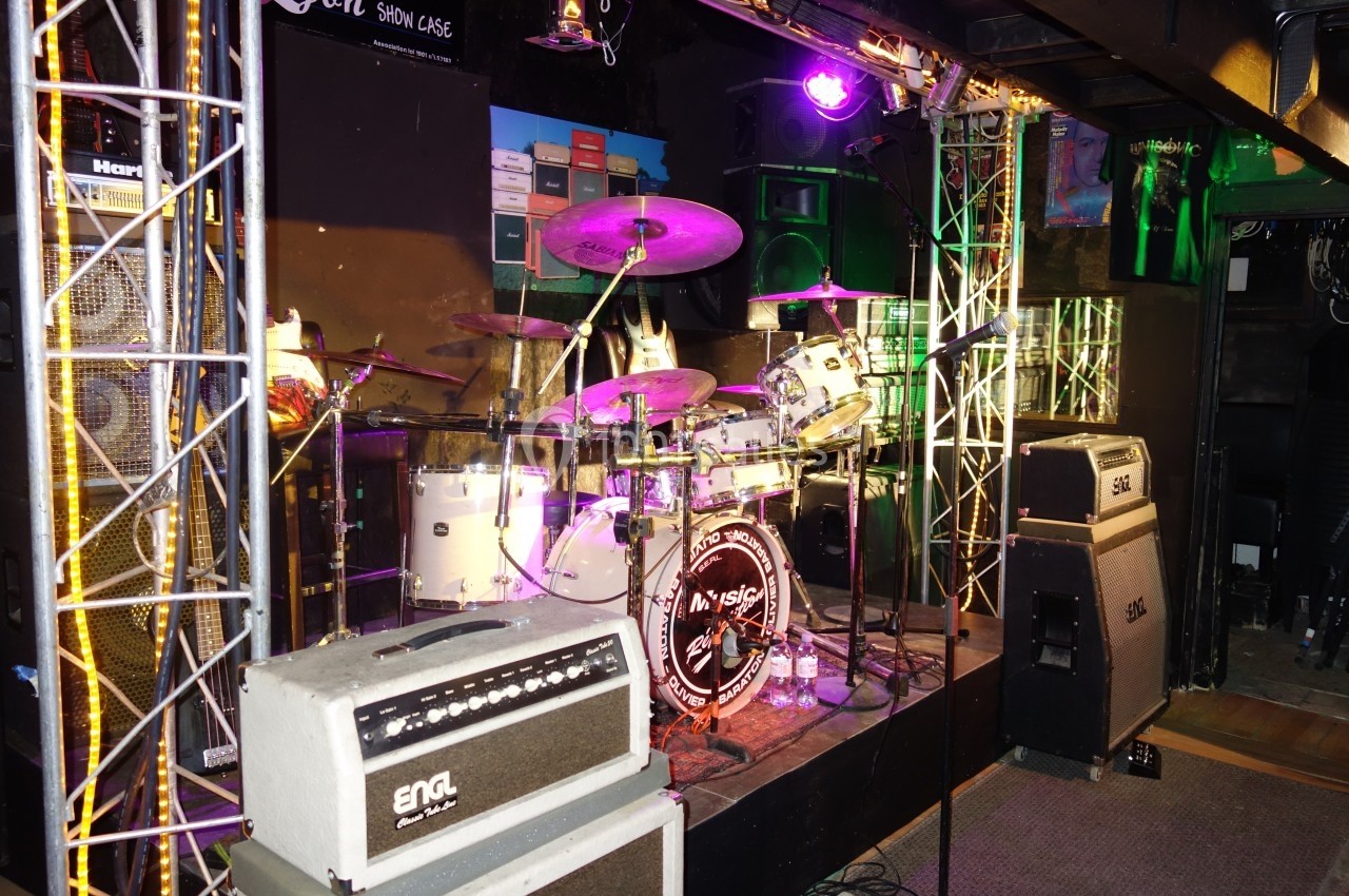 Batterie blanche installée sur une scène éclairée, entourée d'amplificateurs et de structures métalliques.