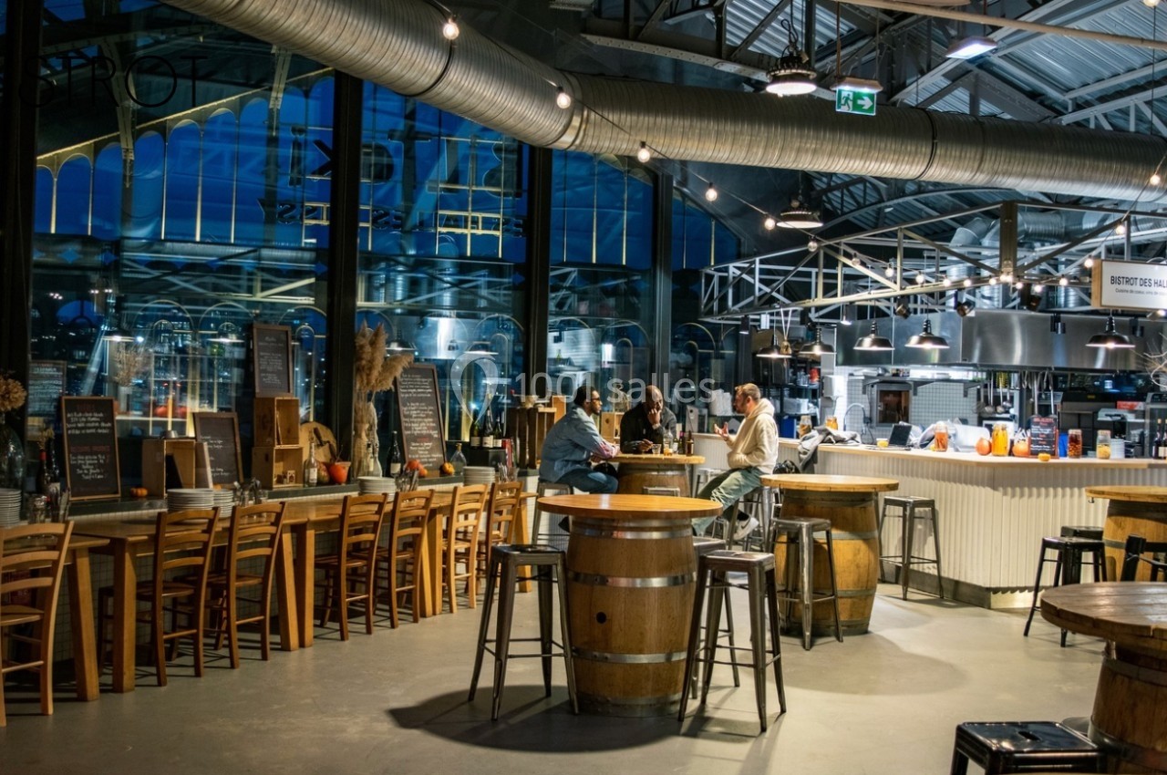 Intérieur d'un restaurant avec tables en bois, bar central éclairé et grandes baies vitrées donnant sur l'extérieur.