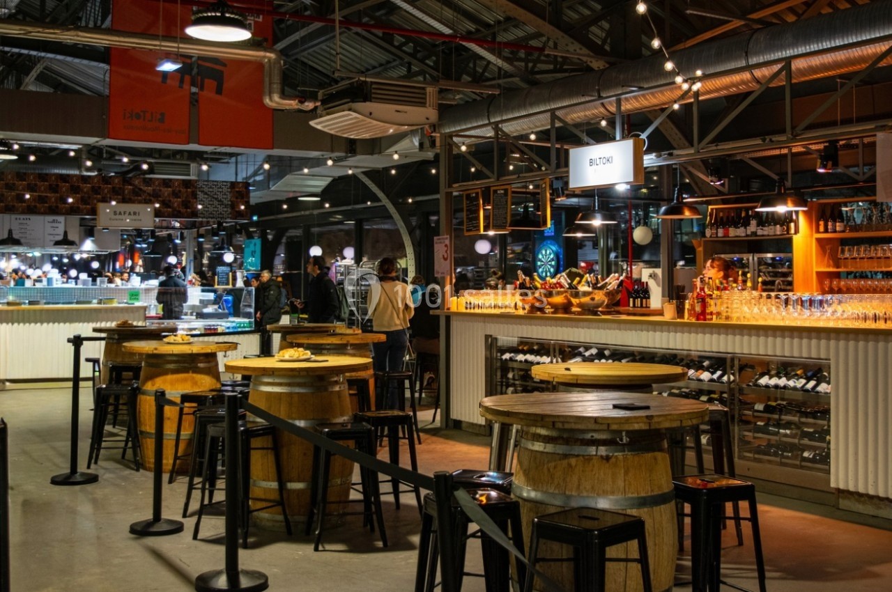 Intérieur d'un marché couvert avec un bar en bois, des tables rondes et des étals éclairés en arrière-plan.