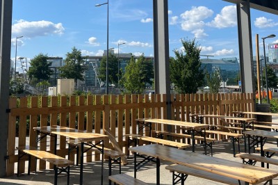Terrasse extérieure avec tables et bancs en bois, entourée d'une clôture, vue sur des bâtiments modernes et un ciel dégagé.