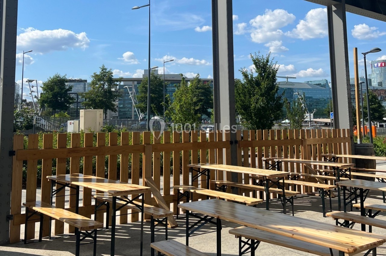 Terrasse extérieure avec tables et bancs en bois, entourée d'une clôture, vue sur des bâtiments modernes et un ciel dégagé.
