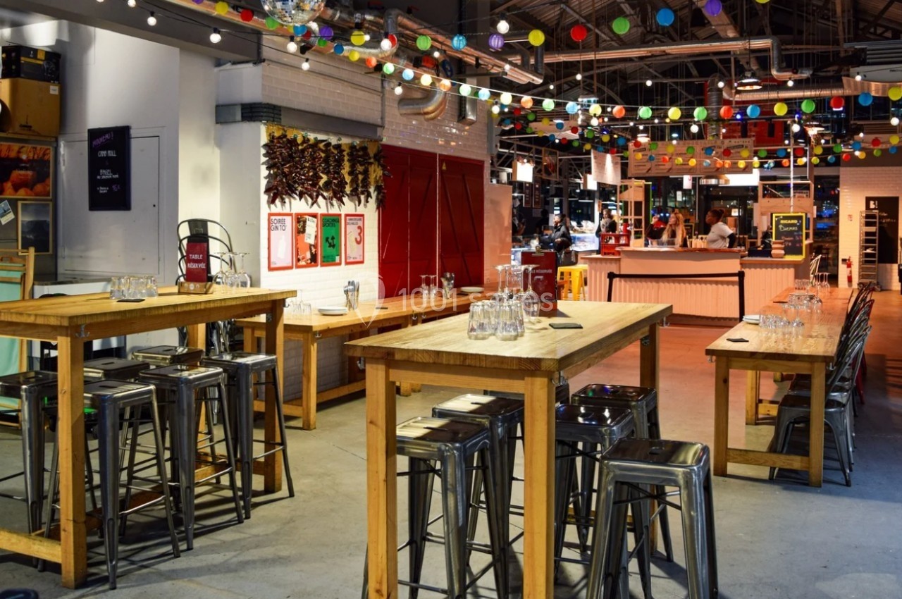 Salle lumineuse avec tables hautes en bois, tabourets métalliques et guirlandes colorées suspendues au plafond.