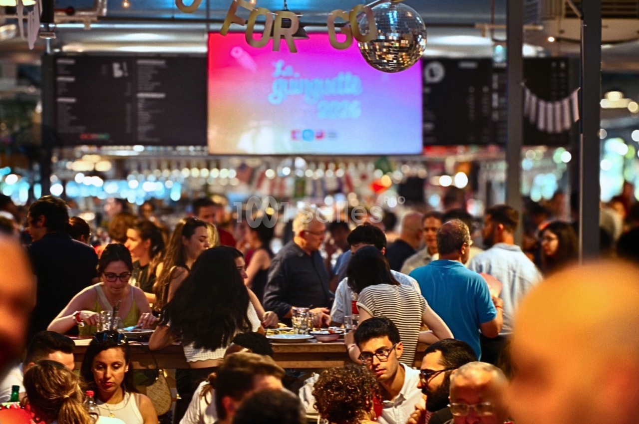 Salle bondée avec des personnes assises à des tables, éclairage chaleureux et écran affichant un événement en arrière-plan.