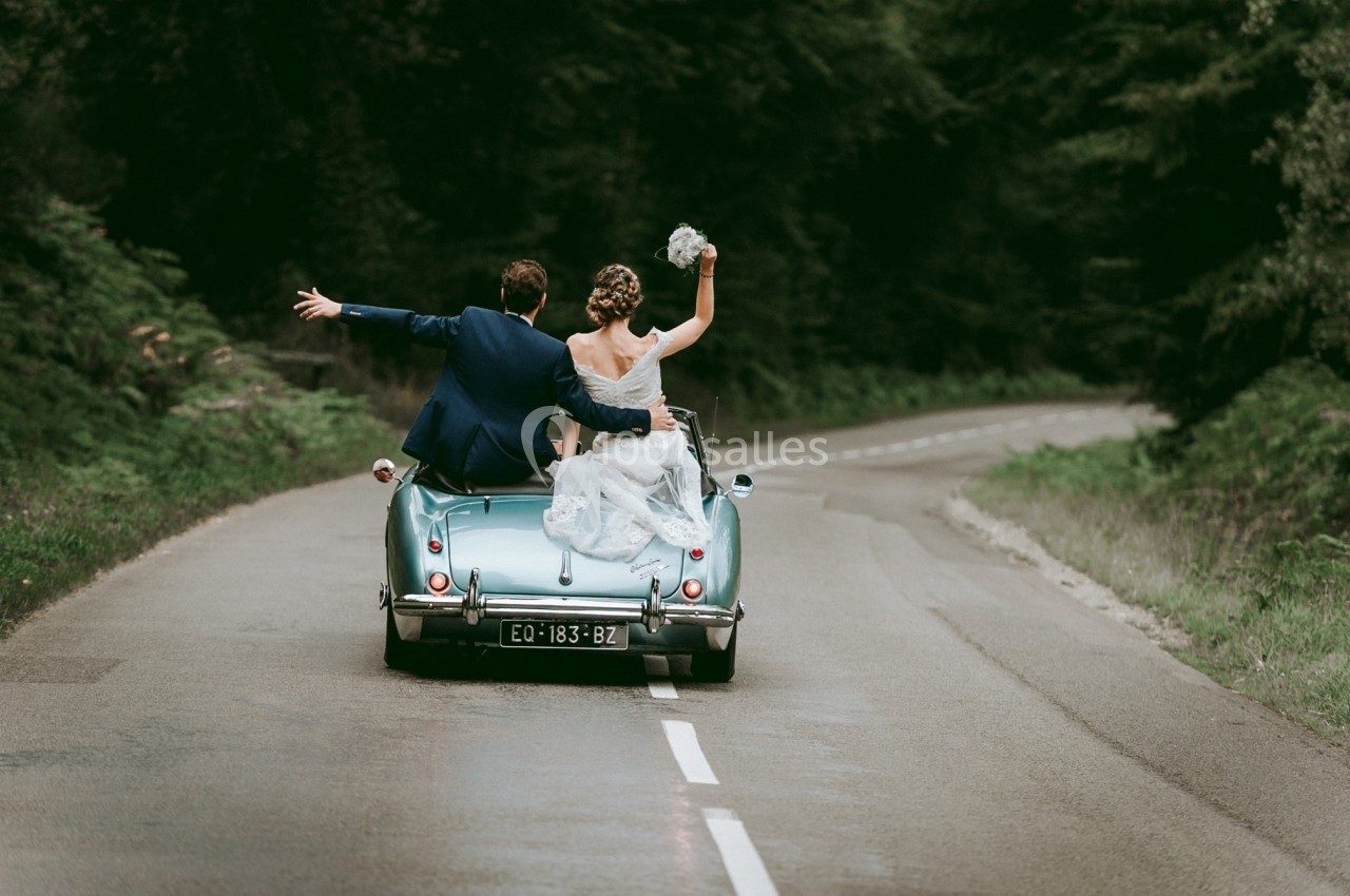 Un couple en tenue de mariage sur une voiture ancienne roule sur une route bordée de forêt.