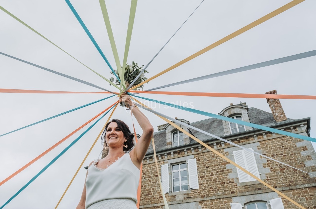 Femme souriante tenant un bouquet au centre de rubans colorés tendus devant un bâtiment en pierre.