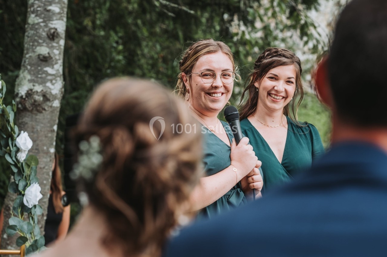 Deux femmes souriantes, l'une tenant un micro, s'expriment devant un petit groupe en extérieur près d'arbres.