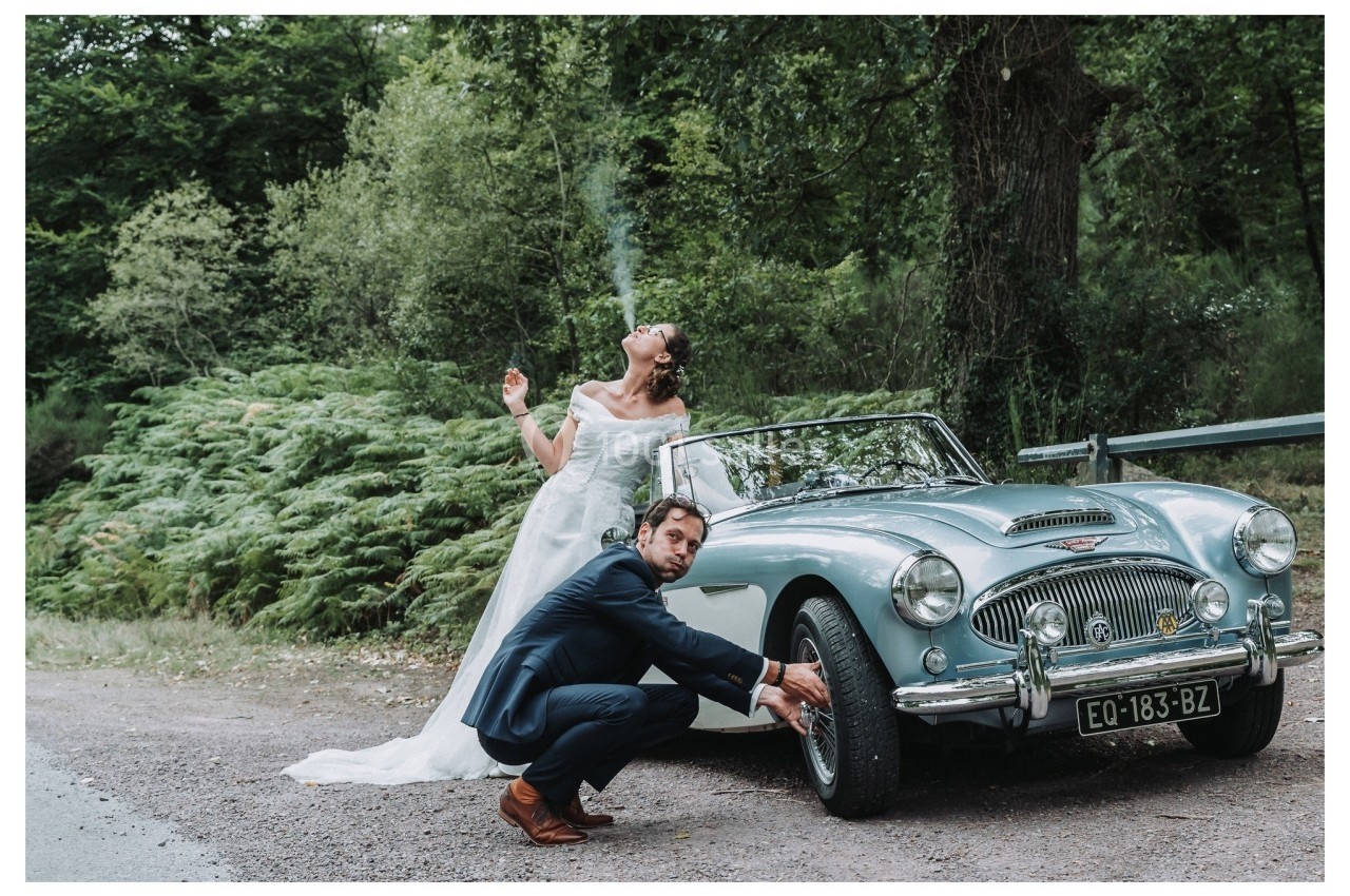 Une mariée fume près d'une voiture ancienne tandis qu'un homme en costume examine une roue sur un chemin forestier.