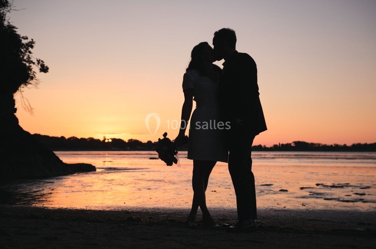 Un couple s'embrasse au bord de l'eau au coucher du soleil, tenant un bouquet de fleurs.
