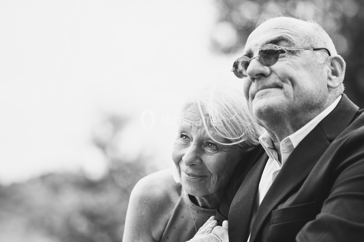 Un couple âgé souriant, assis côte à côte en extérieur, regardant au loin dans un cadre naturel.