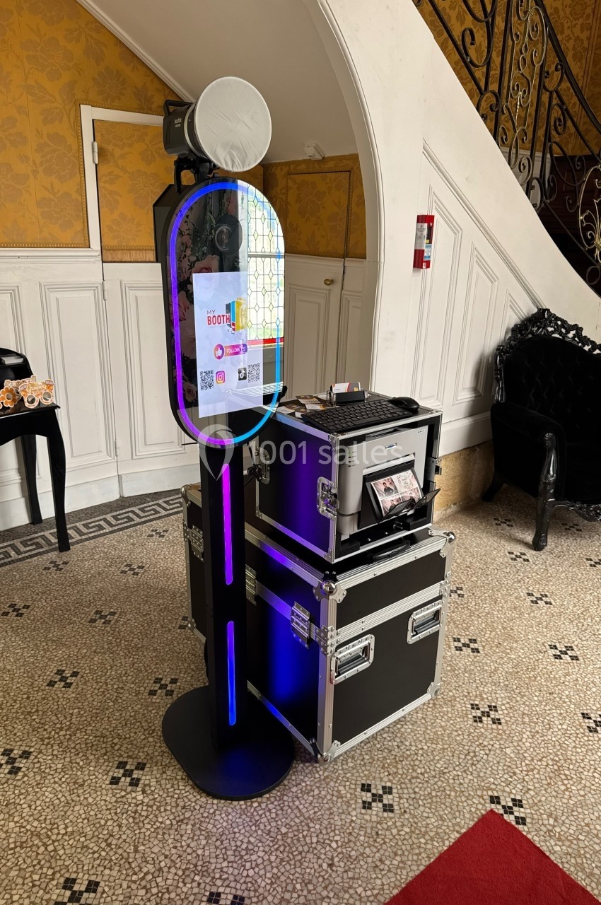 Borne photo interactive avec écran tactile et imprimante, installée dans un hall au sol carrelé et décor classique.