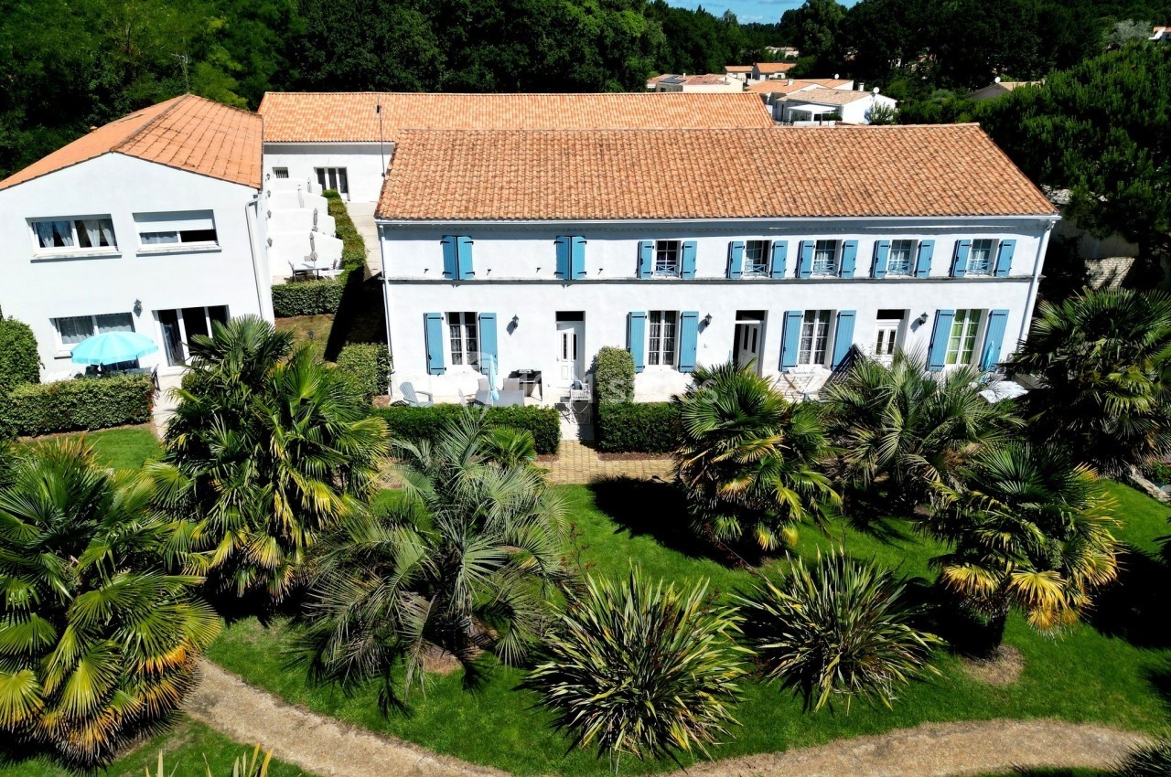 Grande maison blanche aux volets bleus entourée de palmiers et d'un jardin verdoyant sous un ciel ensoleillé.