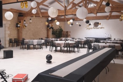 Salle de réception décorée pour un anniversaire, avec tables alignées, guirlandes et lanternes suspendues au plafond en bois.